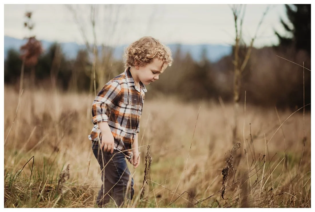 A young boy with blonde curly hair wearing a plaid shirt and jeans, standing in a field of tall, dry grass in Portland, Oregon.