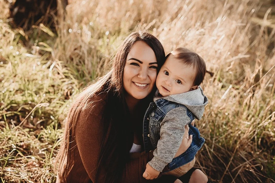 A mother with long dark hair smiling warmly while hugging her young son, who is wearing a denim vest, in a sunlit field with tall dry grass.