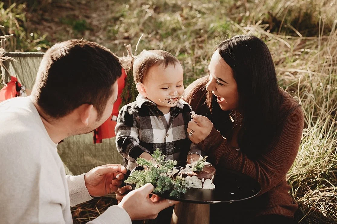 A mother and father laugh and help their young son eat his chocolate-dripped birthday cake during an outdoor cake smash session in a grassy field.