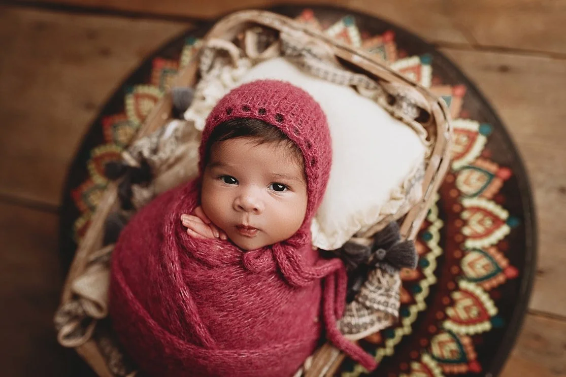 A newborn baby girl with dark hair and brown eyes wrapped in a deep magenta swaddle and matching bonnet, lying in a rustic basket atop a colorful patterned mandala rug.