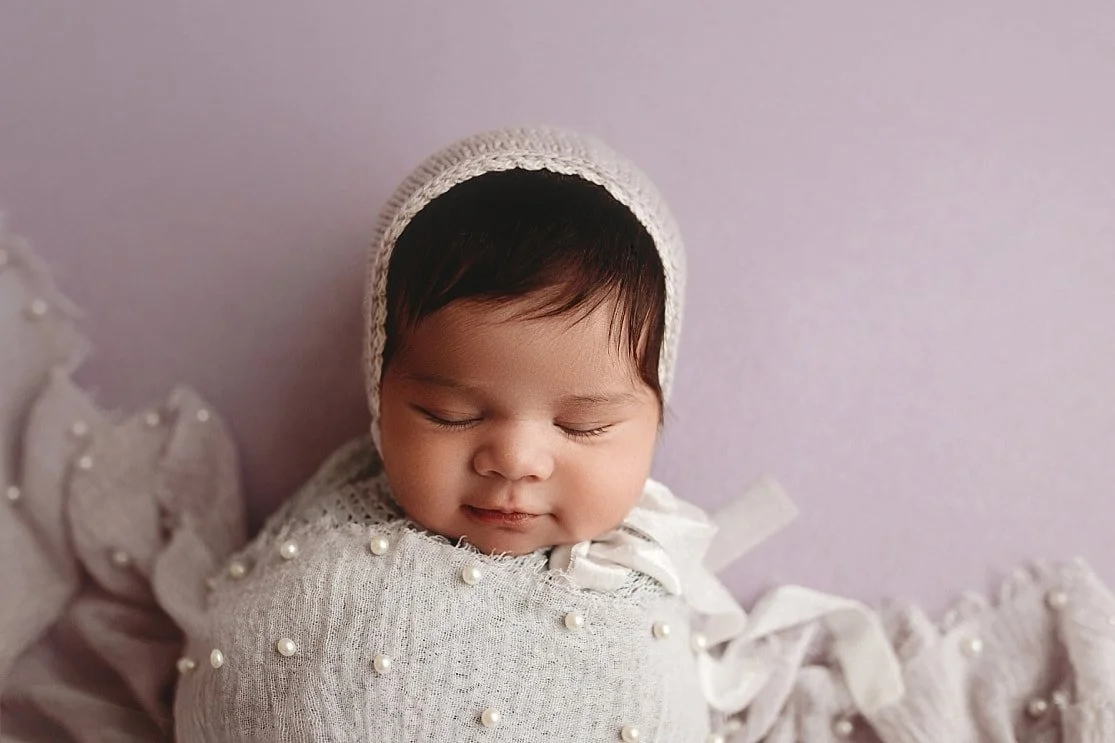 A close-up portrait of a sleeping newborn baby girl with dark hair, wearing a cream knit bonnet and wrapped in a light gray swaddle adorned with small white pearls against a lavender background.