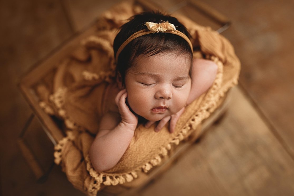 A top-down view of a sleeping newborn baby girl with a dainty bow headband, resting in a wooden crate filled with a mustard yellow fringed blanket.