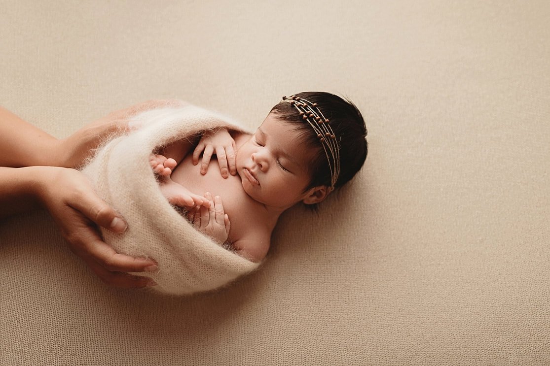 Side-view of a sleeping newborn girl in a cream mohair swaddle and beaded headband, gently cradled by a photographer's hands against a neutral cream background.