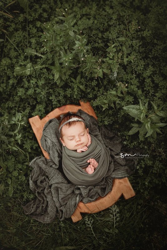 A top-down view of a sleeping newborn baby girl wrapped in a charcoal gray swaddle, resting in a small rustic wooden bed surrounded by lush green garden foliage.