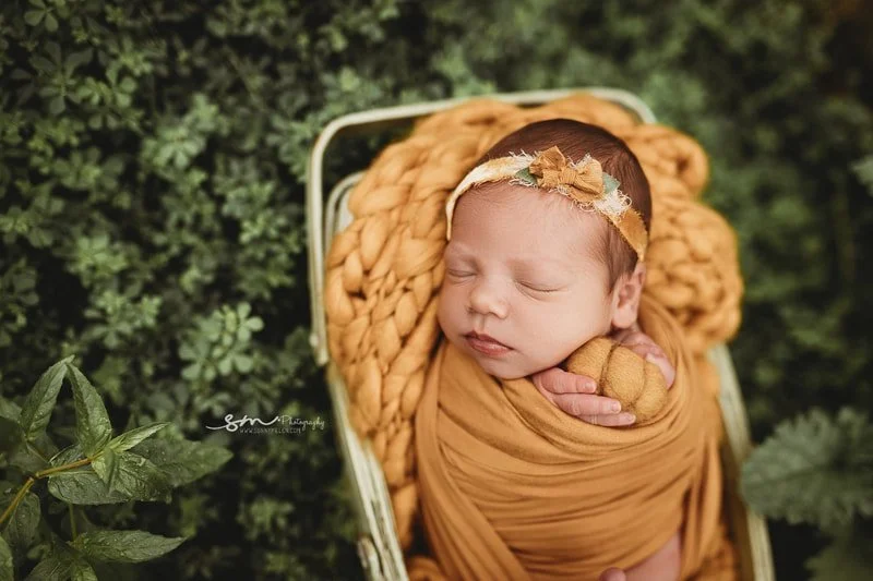 A side-view portrait of a sleeping newborn baby girl wrapped in a mustard yellow swaddle, resting on a chunky knit yellow blanket inside a vintage metal bed prop surrounded by green garden leaves.