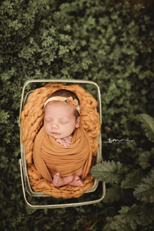 Top-down shot of a sleeping baby girl in a mustard yellow swaddle and chunky knit blanket, resting in a vintage metal bed surrounded by lush green foliage.