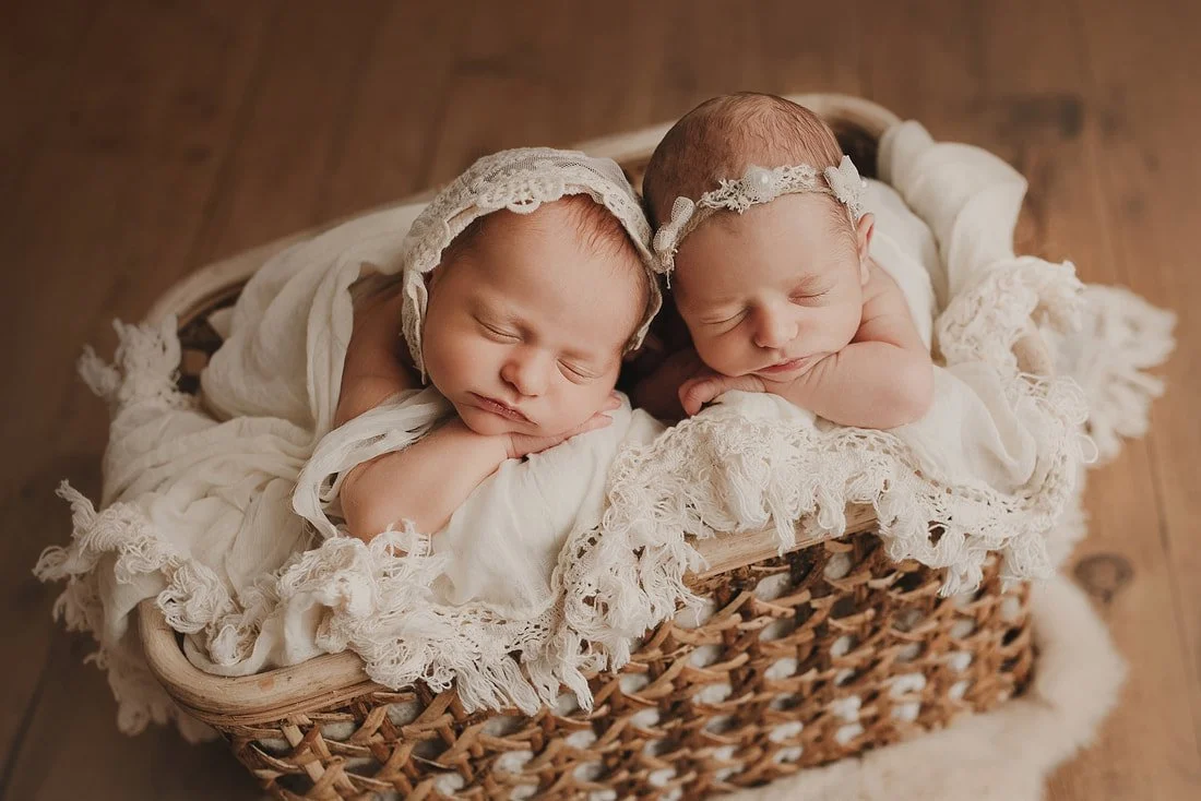 Two sleeping newborn twins posed together in a rustic woven basket; one wears a delicate lace bonnet and the other a floral headband, both resting on soft cream linens.