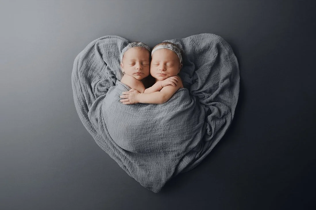 This artistic newborn twin portrait utilizes a custom-sculpted heart-shaped wrap to symbolize the love and connection between siblings in Portland, Oregon . The twins are expertly posed in a gentle embrace, tucked into a rich slate gray textured fabric that blends seamlessly with the minimalist studio backdrop. Both infants are styled with delicate, neutral-toned headbands, maintaining a sophisticated and modern aesthetic. This image is a premier example of the creative vision and technical posing skills provided by the best twin photographers in the area , offering families a unique and sentimental masterpiece to document their children’s earliest days.