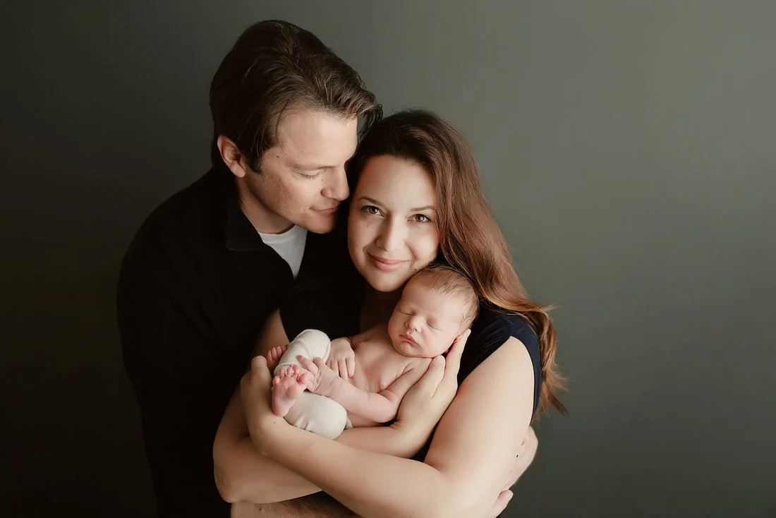 A mother and father in dark clothing lovingly cradling their sleeping newborn baby against a dark olive green studio backdrop.