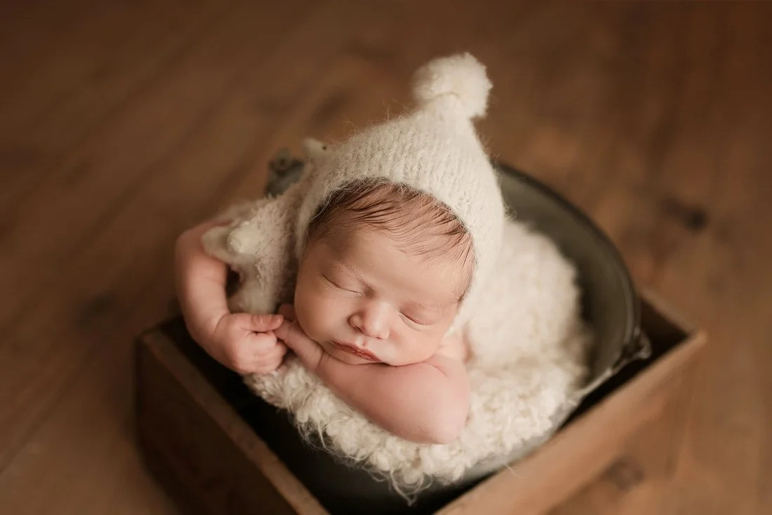 A top-down view of a sleeping newborn baby boy wearing a cream knit pointed sleepy cap, nestled in a rustic bucket with a plush white rug inside a wooden crate.