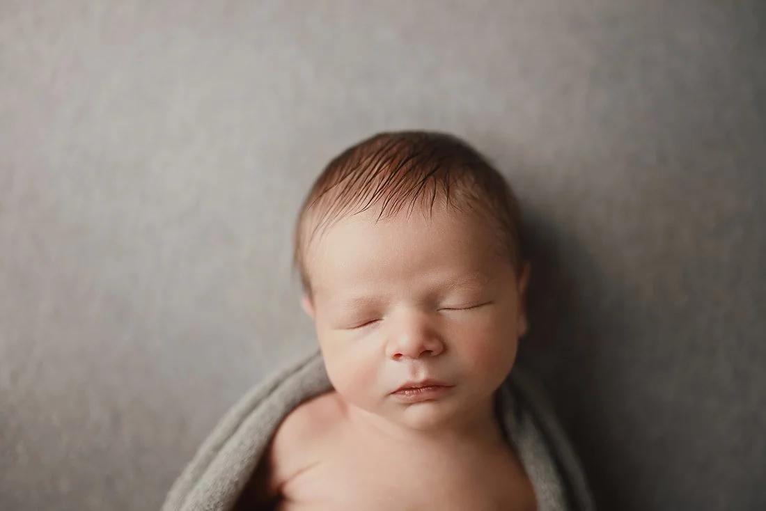 A close-up, top-down portrait of a sleeping newborn baby with soft brown hair, wrapped in a light gray swaddle against a textured gray studio backdrop.