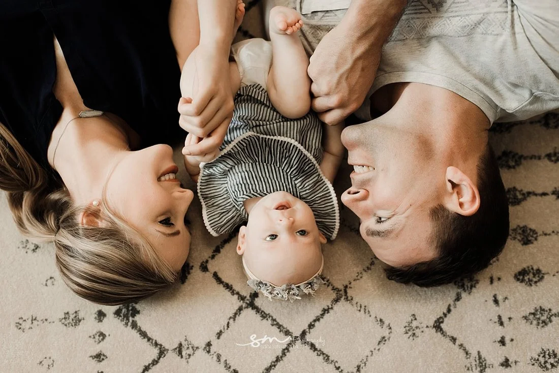 An overhead view of a mother and father lying on a cream geometric rug, smiling at their baby girl who is lying between them wearing a striped romper and a floral headband.