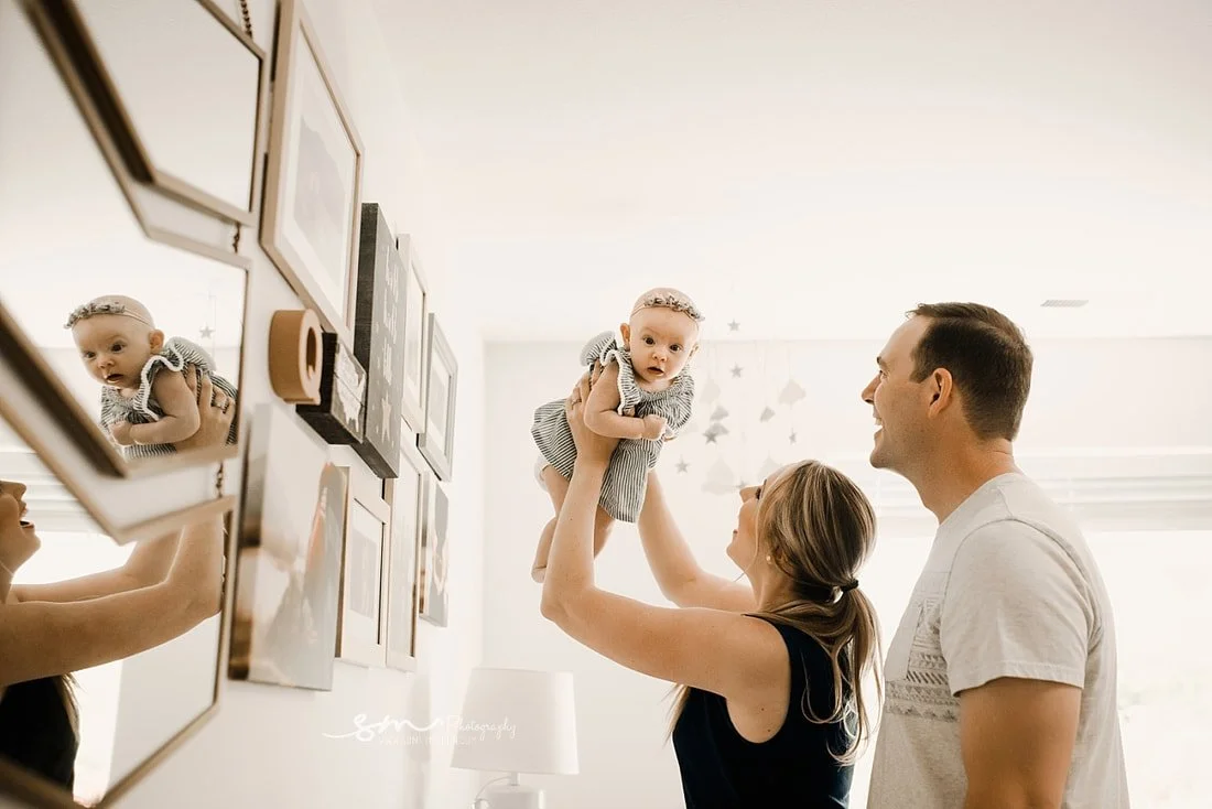 A mother lifting her baby girl high in the air while the father smiles beside them, standing in front of a modern gallery wall with multiple framed photos and mirrors in a bright nursery.