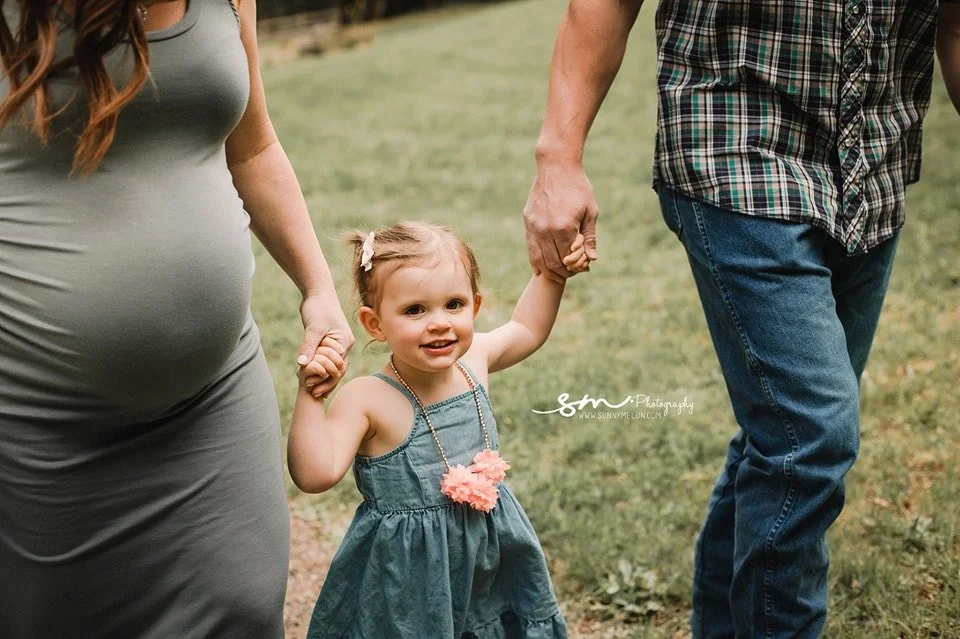 A close-up of a young girl in a blue dress with pink floral tassels holding hands with her pregnant mother and her father while walking through a green field.