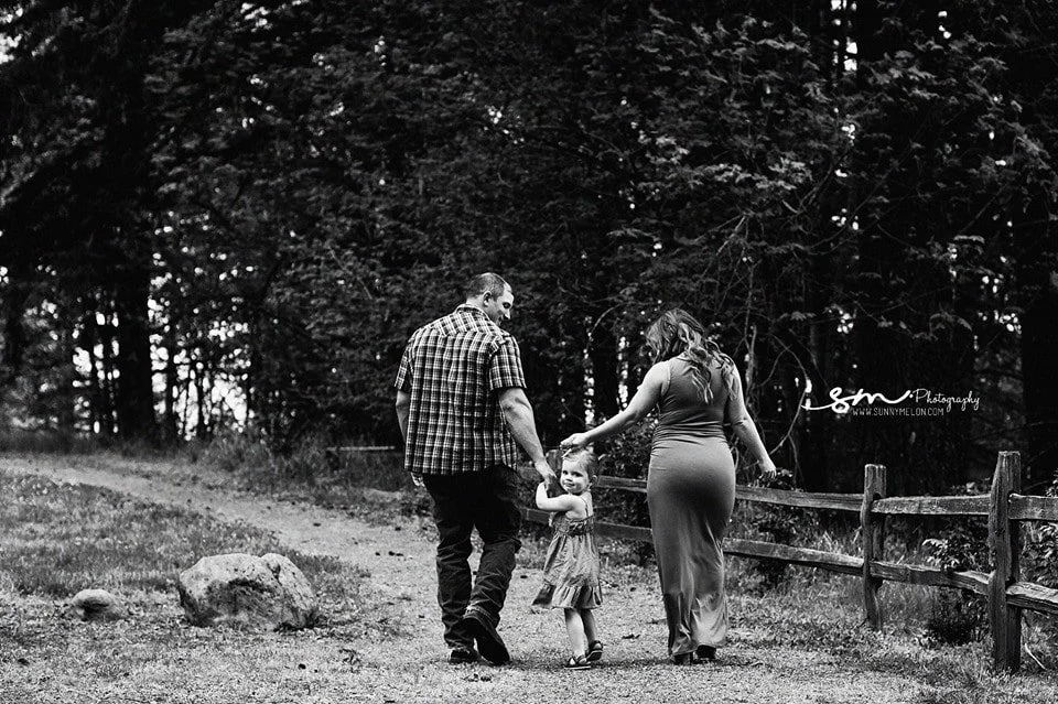 A black and white photograph of a pregnant mother in a fitted dress and a father in a plaid shirt walking away on a forest path while holding the hands of their young daughter.