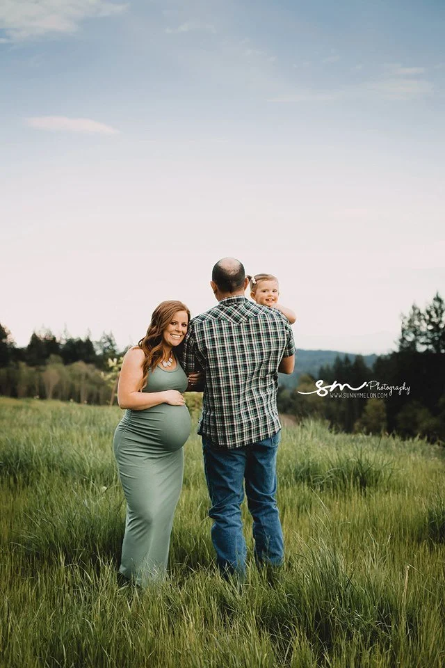 A pregnant woman in a sage green dress and a man in a plaid shirt holding a toddler, standing in a grassy field with a forest and mountain backdrop.
