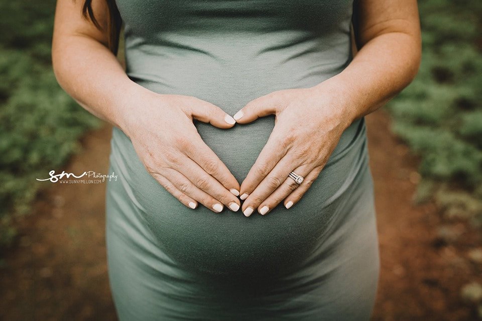A close-up of a pregnant woman's hands forming a heart shape over her baby bump while wearing a sage green fitted dress in an outdoor setting.