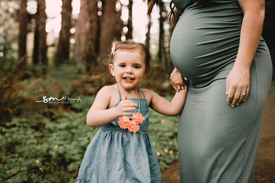 A close-up of a smiling toddler girl in a blue dress standing next to her pregnant mother in a green fitted dress in a sunlit forest.