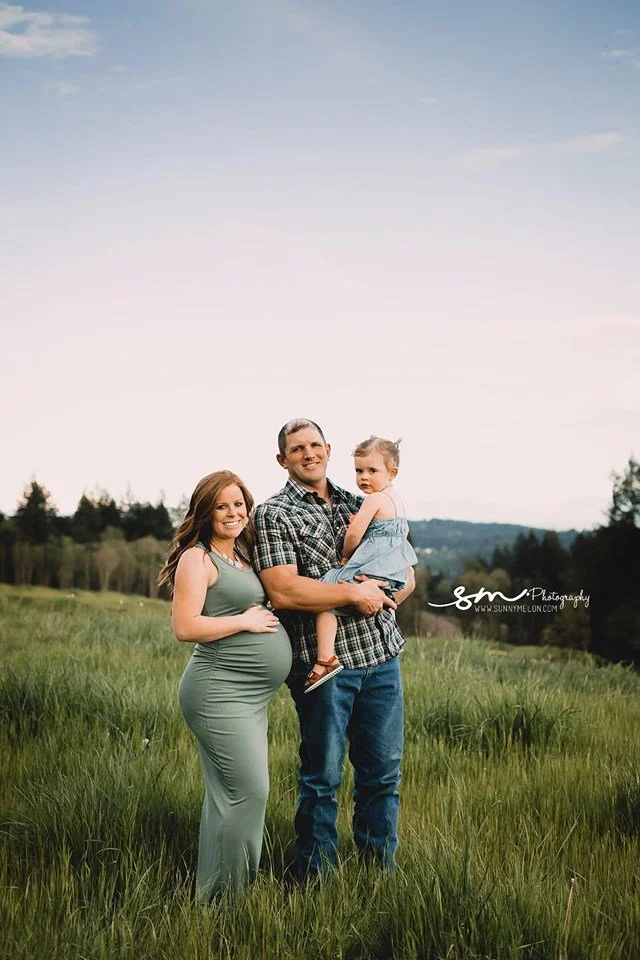 A pregnant woman in a sage green dress standing next to a man in a plaid shirt holding a toddler, all posing in a grassy field with a forest background under a soft sky.