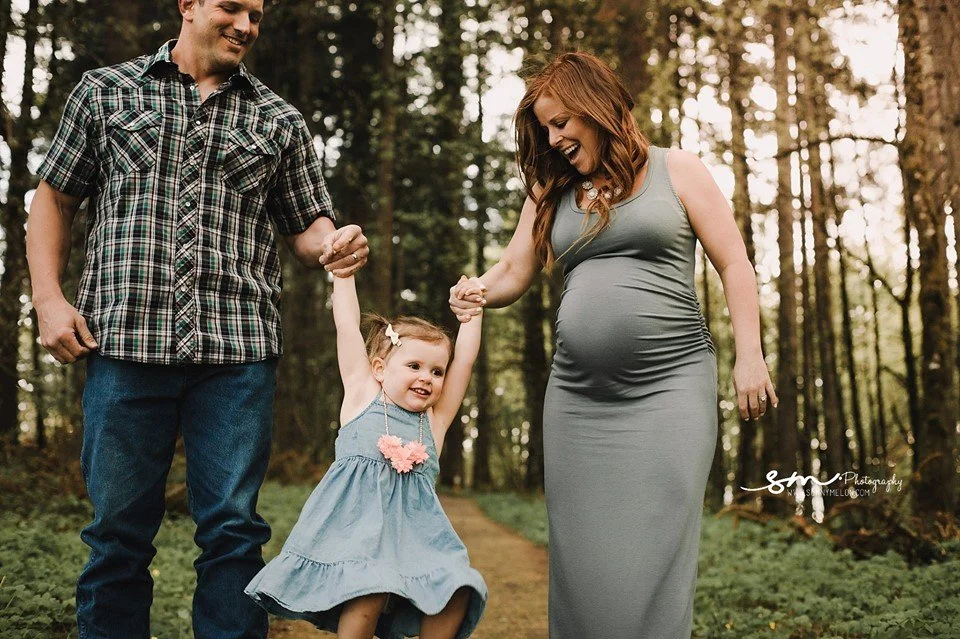 A happy pregnant mother in a sage green dress and a father in a plaid shirt lifting their toddler daughter into the air while walking along a wooded path in a forest.