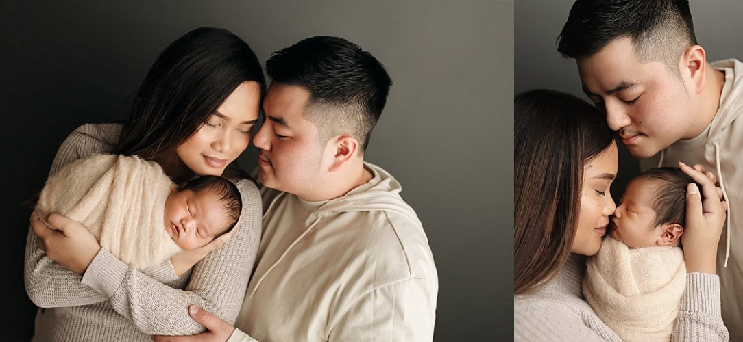 Professional studio portrait of a mother and father cradling their sleeping newborn baby against a dark background.