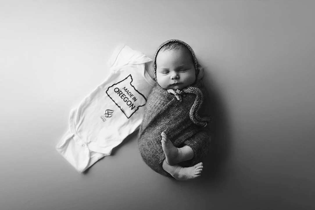 A fine-art black and white overhead portrait of a sleeping newborn baby wrapped snugly next to a white onesie featuring a "Made in Oregon" graphic.