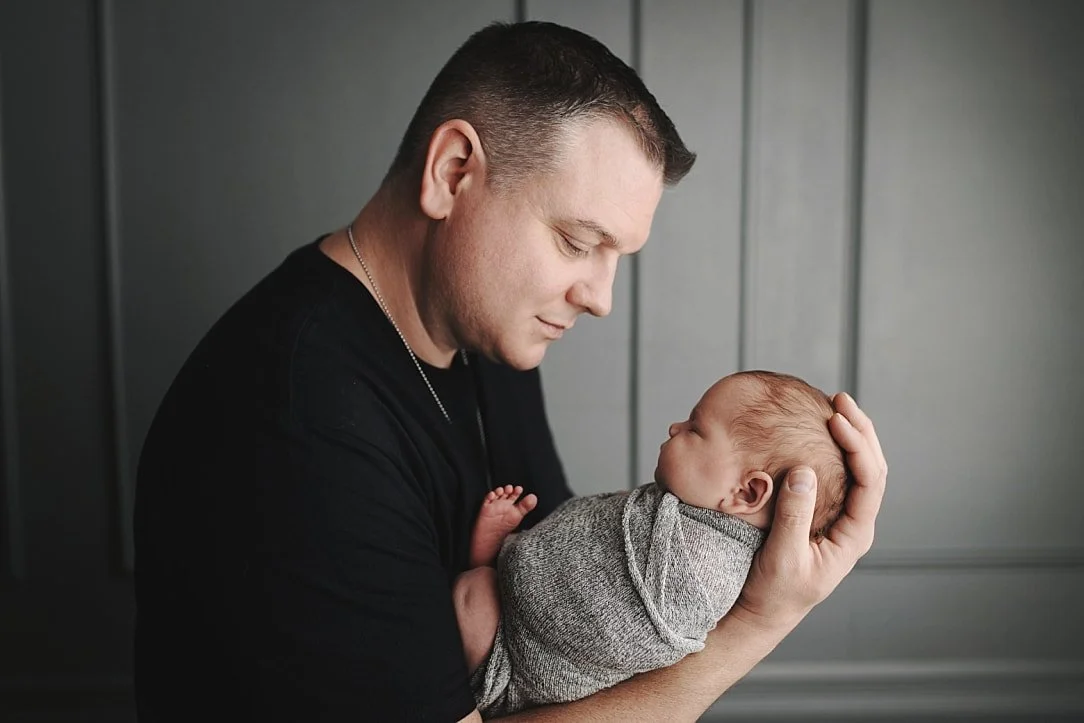 A classic studio portrait of a father dressed in a black t-shirt, viewed from the side as he lovingly holds his sleeping, swaddled newborn baby in his arms against a grey paneled wall background.