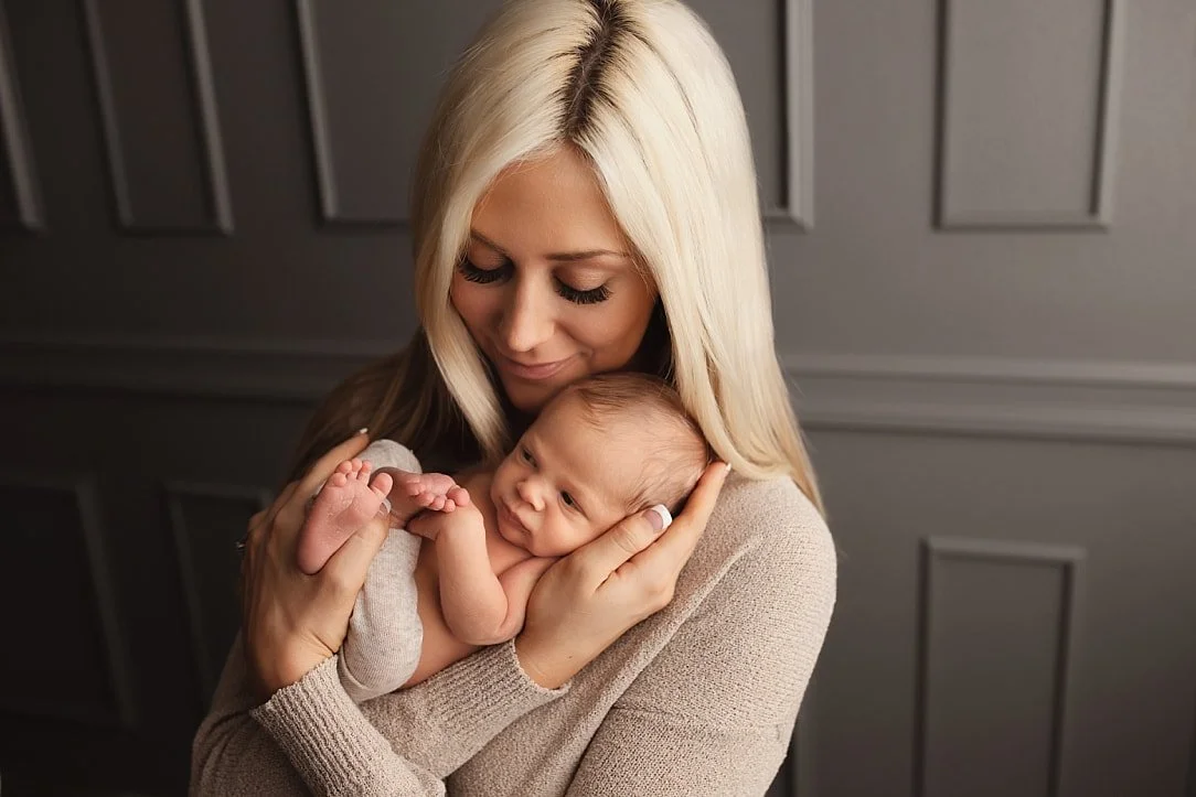 A close-up studio portrait of a blonde mother smiling down lovingly while cradling her newborn baby in her arms against a classic grey paneled wall background.