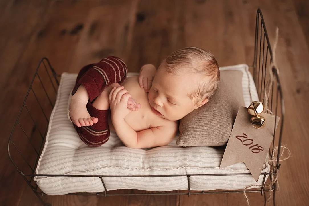 A sleeping newborn boy curled on a miniature vintage metal bed prop. He wears burgundy striped leg warmers and rests his head on a small pillow, alongside a rustic paper tag marked "2018" with decorative bells.