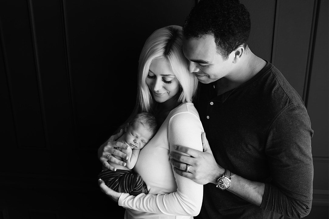 A classic black and white studio portrait of a mother and father holding their sleeping newborn baby tightly, posed affectionately against a dark paneled wall.