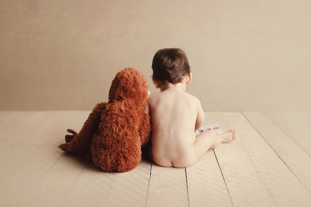 A sweet, minimalist back-view portrait of a shirtless toddler sitting on a light-colored wooden floor next to a plush brown orangutan. The duo is posed against a warm neutral background, capturing a quiet moment of childhood wonder.