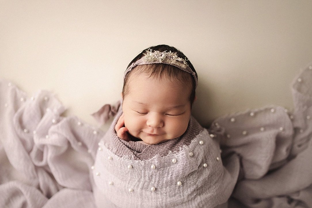 A serene studio portrait of a smiling newborn baby girl wearing a delicate floral headband. She is swaddled in a lavender-toned wrap embellished with pearls, set against a clean, neutral cream background.