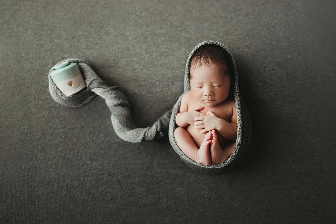 A poignant memorial portrait of a newborn twin sleeping peacefully in a gray knit wrap. The wrap extends gracefully across a dark, textured background to encircle a small keepsake urn, symbolizing the baby's twin in a beautiful, artistic tribute.