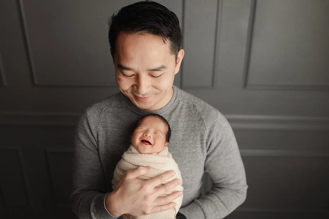 A touching professional studio portrait of a father in a grey sweater looking down lovingly at his swaddled newborn baby, who is captured with a joyful smile. The pair is posed against a classic grey paneled wall backdrop.