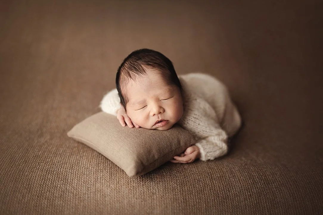 A peaceful newborn portrait featuring a sleeping baby in a cream-colored knit sweater. The infant is resting their head on a tiny tan pillow against a warm, textured brown studio backdrop.