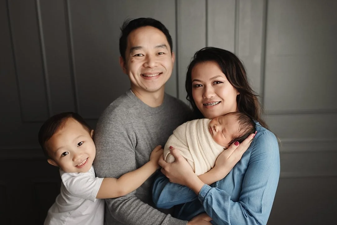 A warm, professional studio portrait of a family of four. A mother in blue holds a swaddled newborn, while the father in a grey sweater smiles alongside them as their young son hugs his arm, all posed against a classic grey paneled wall.