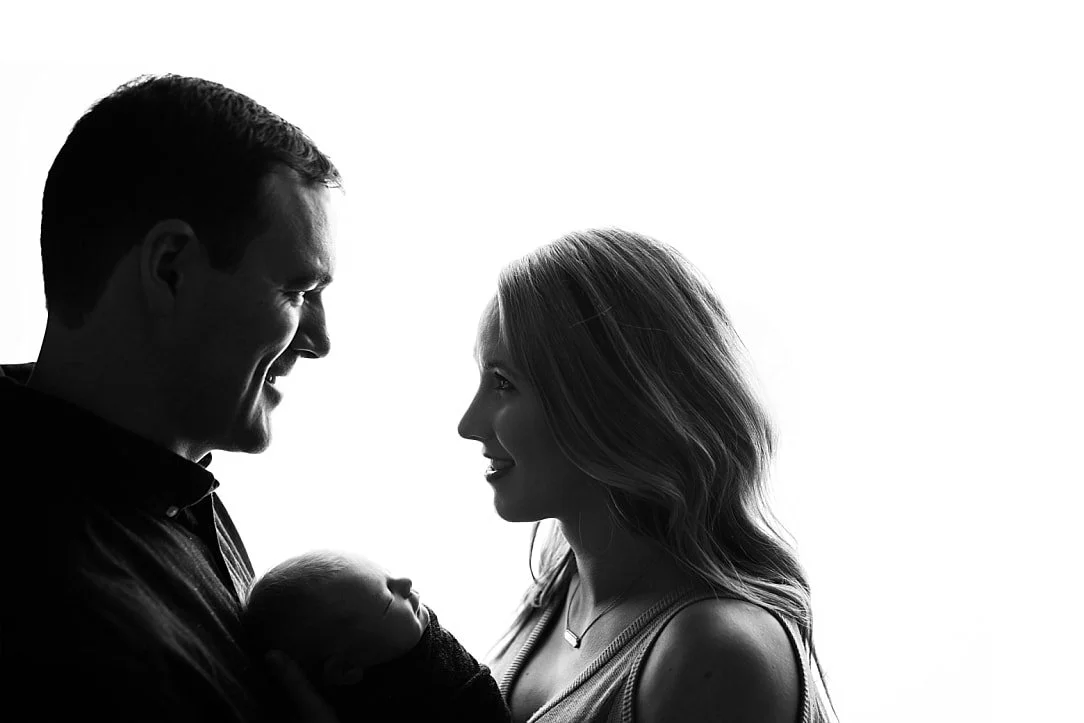 A high-contrast, black and white silhouette portrait of a smiling mother and father looking at each other while cradling their newborn baby against a bright white background.