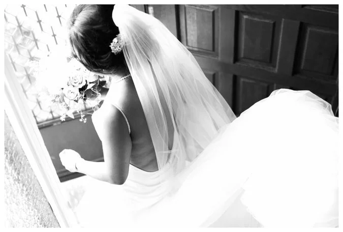 A high-angle black and white photograph of a bride from behind, showcasing a backless wedding dress and a long, airy veil as she stands near a door.