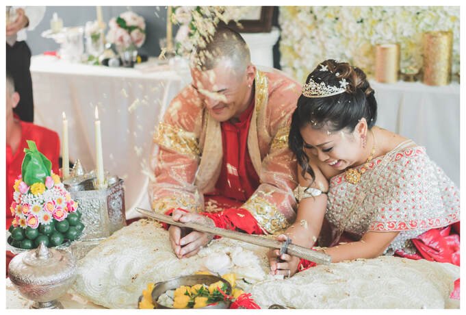 A Cambodian bride and groom in ornate pink and gold traditional silk garments smiling as guests toss seeds or flower petals during the hair-cutting ritual at their wedding.