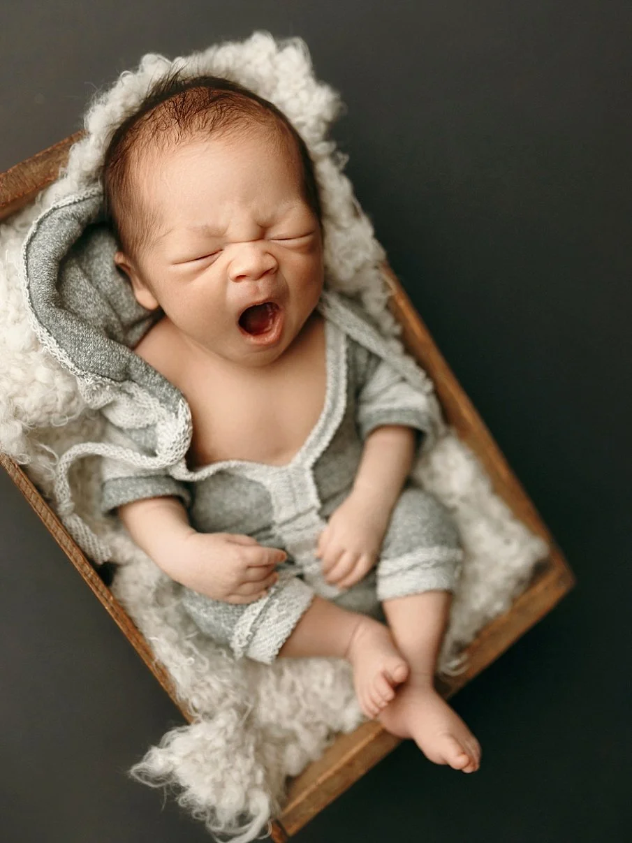 A charming and candid newborn photograph capturing an infant mid-yawn. The baby is dressed in a soft, grey textured romper with white trim and is lying on a plush, white wool-like blanket inside a vintage wooden crate. The crate is positioned at an a