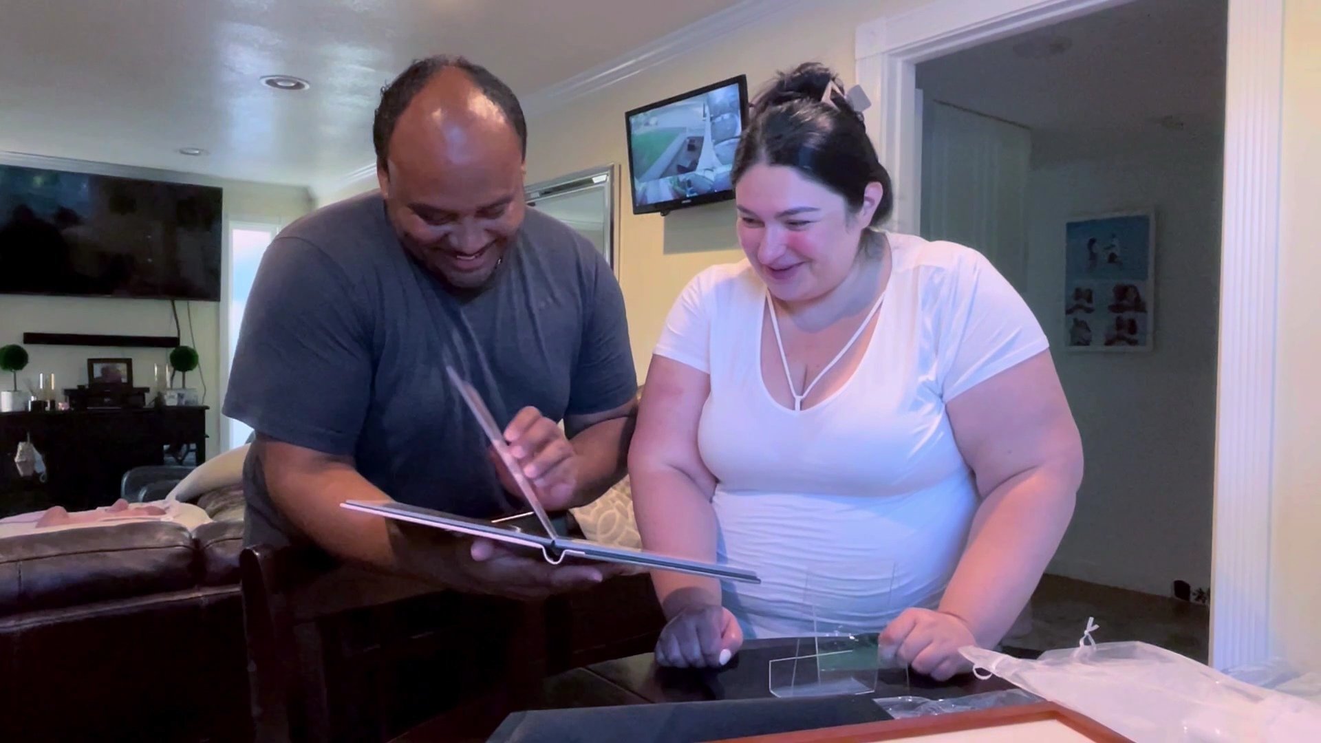 A heartwarming indoor scene featuring a man and woman smiling joyfully as they flip through a high-quality, professional photo album together. The couple is leaning over a table in a home setting, illuminated by soft indoor lighting, capturing the au