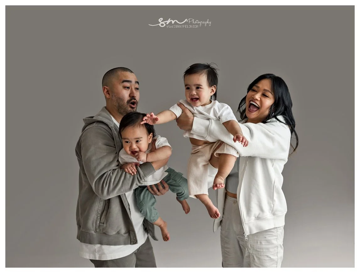 A candid studio portrait against a grey background showing a mother and father holding their twin toddlers; the mother laughs while lifting one twin mid-air, and the father holds the other twin reaching out.