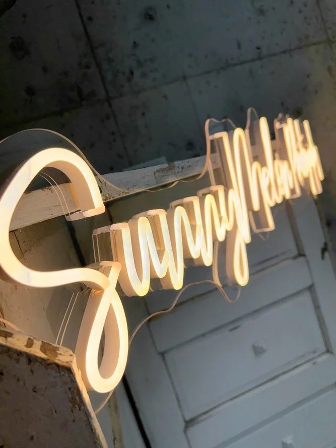A glowing warm white neon sign spelling out "Sunnymelon Photography" in a script font, mounted on a rustic white brick wall in a professional photo studio.