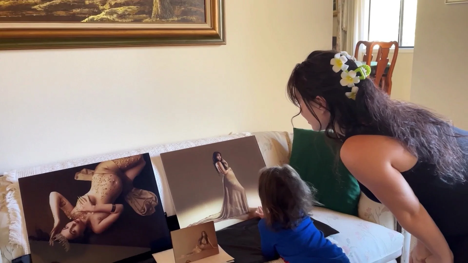 This intimate lifestyle photograph depicts a mother and her young child bonding over professional maternity portraits displayed on a sofa. The high-contrast, artistic prints showcase the beauty of pregnancy, while the mother’s and child’s focused att
