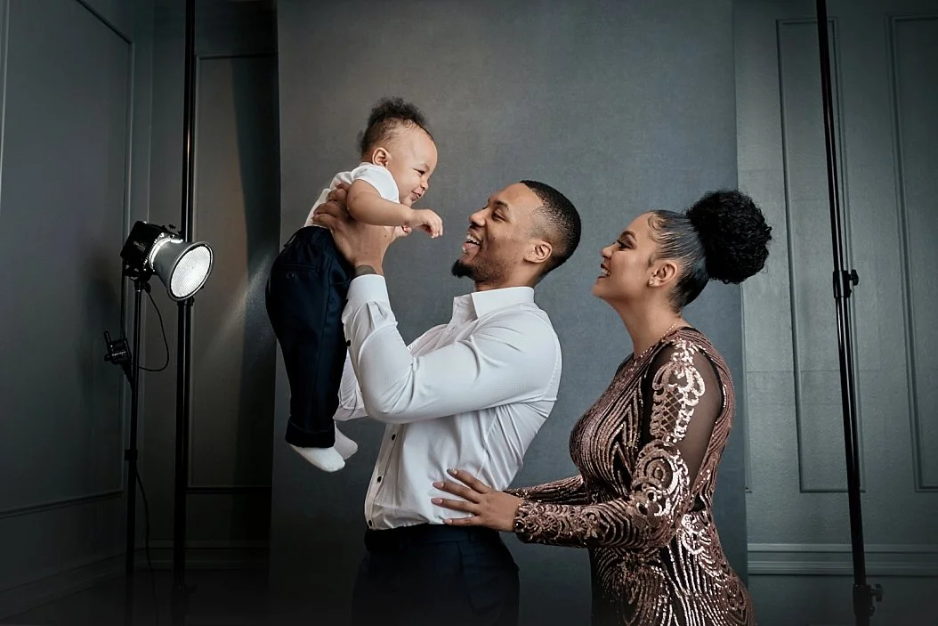This high-end editorial family portrait captures a joyful moment between NBA superstar Damian Lillard, his partner, and their infant. The image features professional studio elements, including a clean grey backdrop and visible lighting equipment, whi