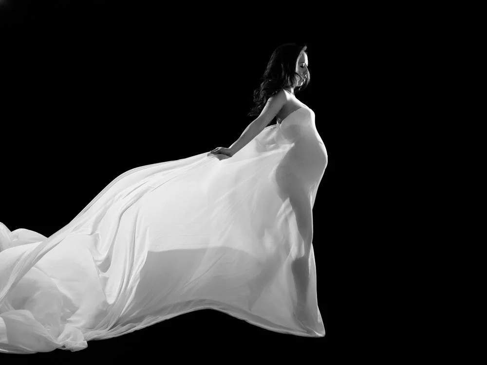 Sunnymelon Photography dramatic black and white fine art maternity portrait. An expectant mother is draped in a flowing white sheer fabric gown against a black background, with her pregnant belly highlighted.