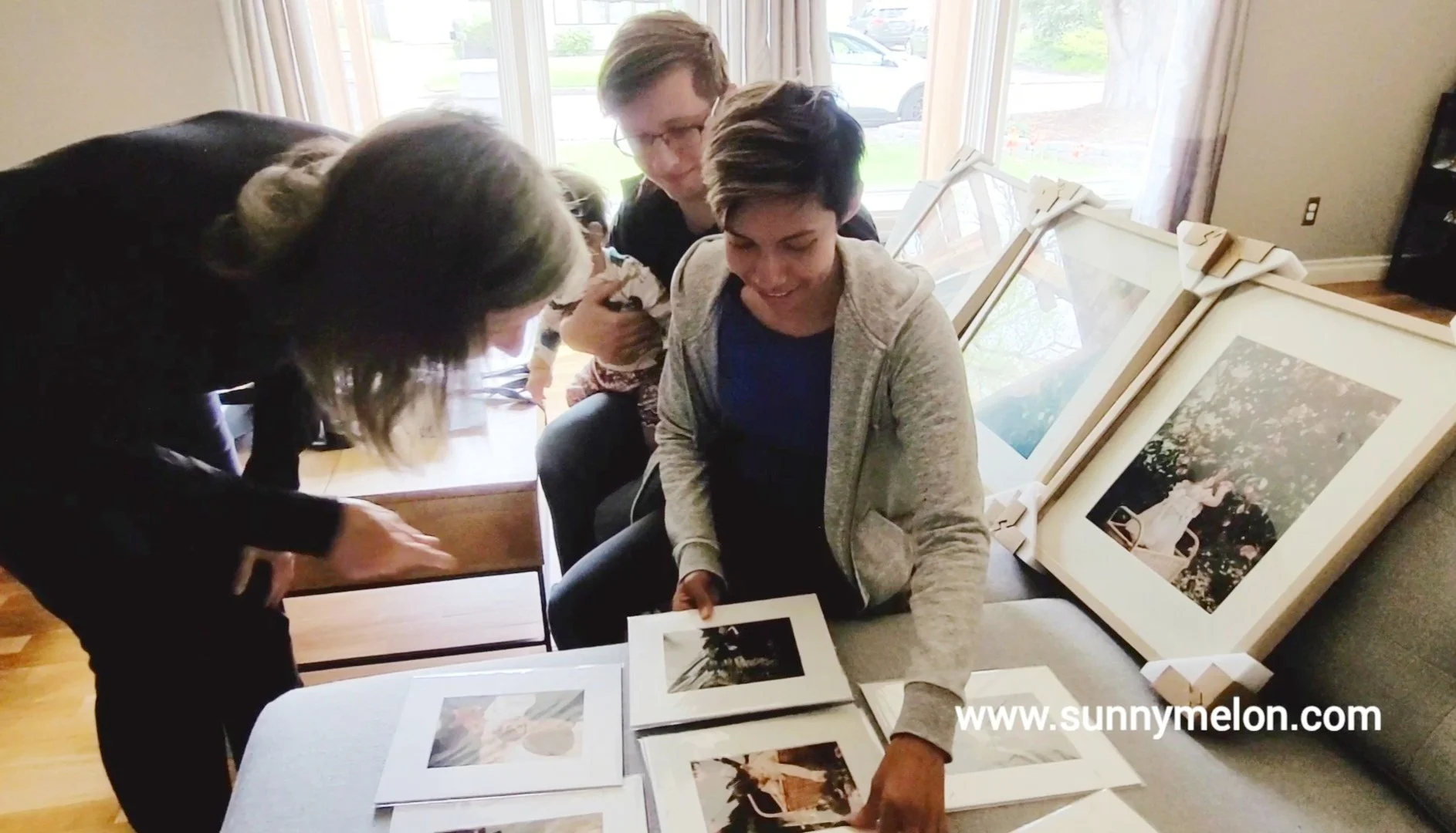 This candid lifestyle image captures a family’s genuine delight during an in-home photography reveal. The scene shows two women and a young child interacting with a variety of high-end matted prints and large-format framed portraits, illustrating the