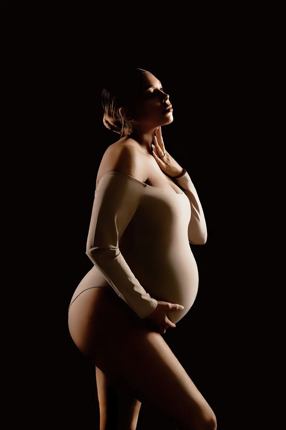 Fine-art studio maternity portrait capturing a pregnant woman's silhouette in a beige bodysuit against a black backdrop in West Linn, Oregon.