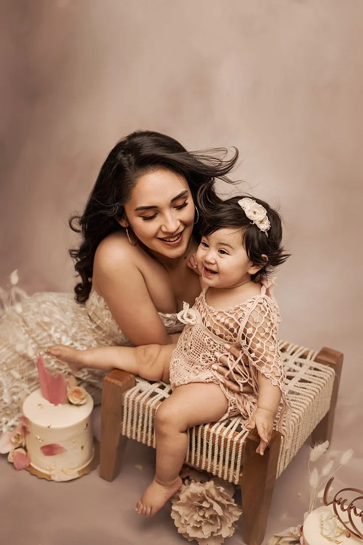 A mother with long dark hair smiles at her laughing one-year-old daughter during a cake smash session. The baby wears a pink crochet romper and floral headband, sitting on a small wooden bench next to a birthday cake in a warm-toned studio.