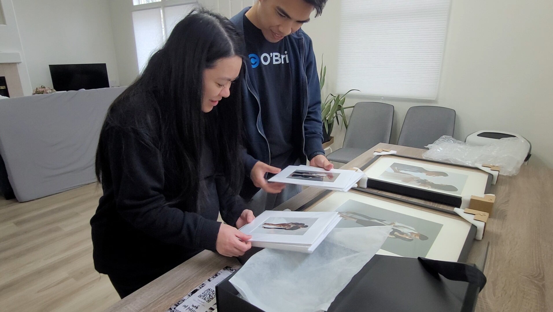 A candid, indoor shot of a man and a pregnant woman smiling while reviewing high-quality matted prints and large framed portraits from their recent photography session.